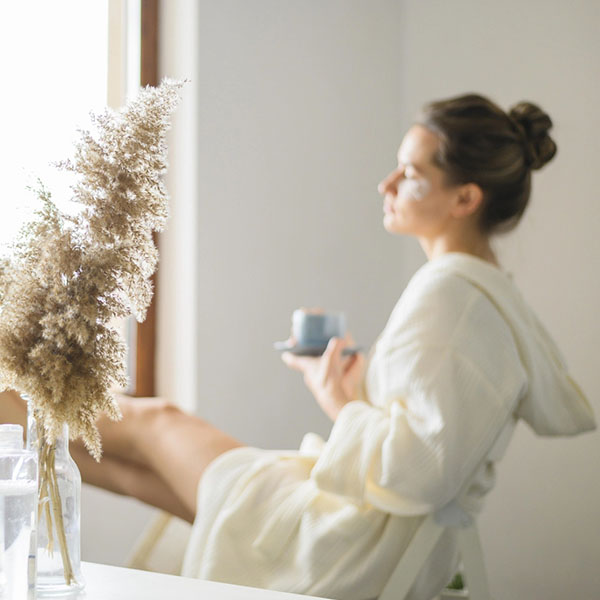Frau im Bademantel sitzt entspannt am Fenster und trinkt Kaffee